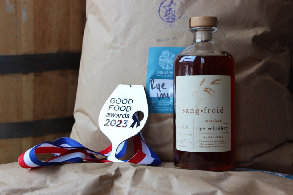 A bottle of Sangfroid rye whiskey sits beside a Good Food Awards 2023 medal, a blue ribbon, and a brown paper bag labeled “Rye Malt.” The scene is set against a wooden barrel background.