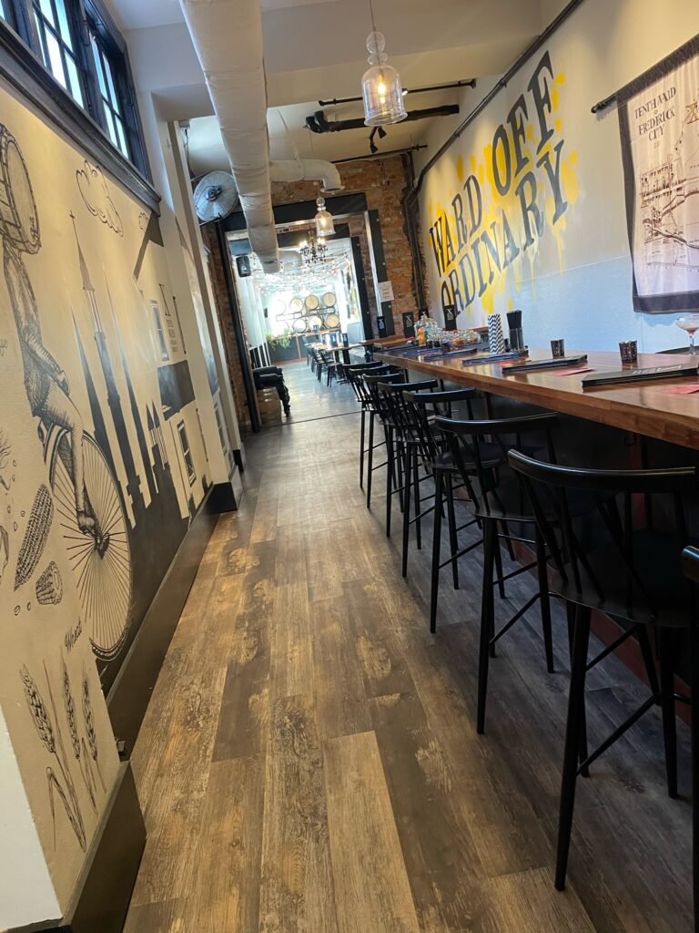 A modern, cozy restaurant with wooden floors, high tables and chairs, mural art on one wall, and “WARD OFF ORDINARY” painted in yellow letters on the opposite wall.