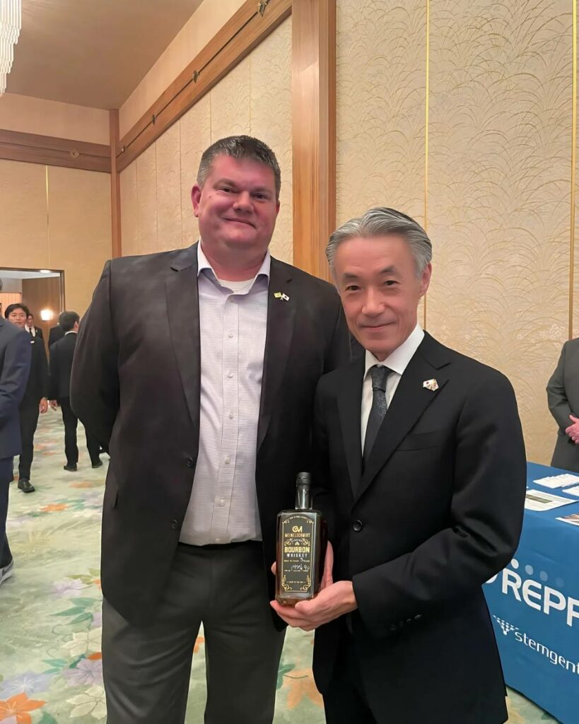 Two men in suits stand indoors at an event. One man holds a bottle with a black and gold label. A blue table with white text and other people are visible in the background.