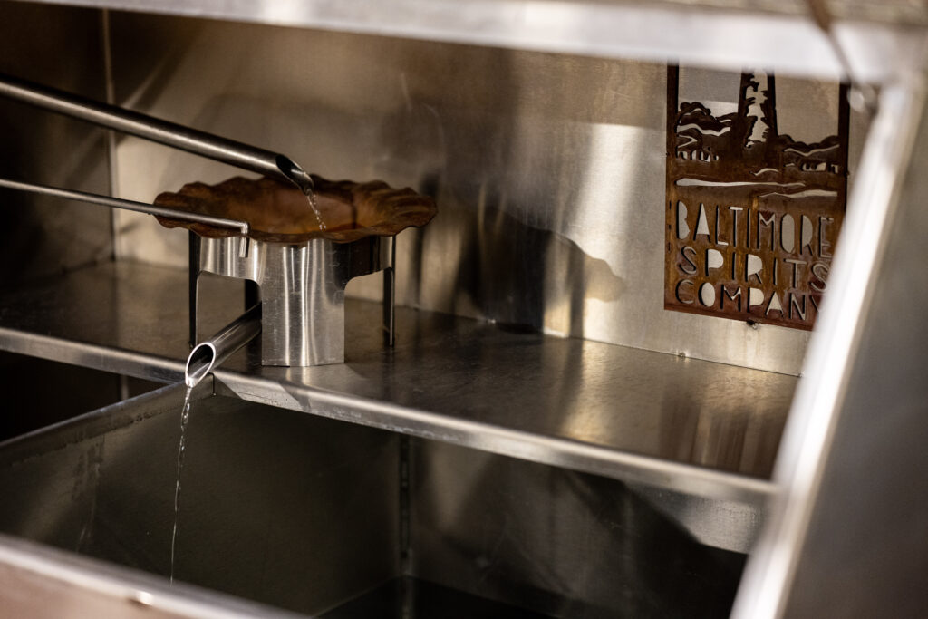 Metal tongs hold a brown leaf under a thin stream of liquid inside a stainless steel distillation setup with a “Baltimore Spirits Company” logo cut into the metal wall.