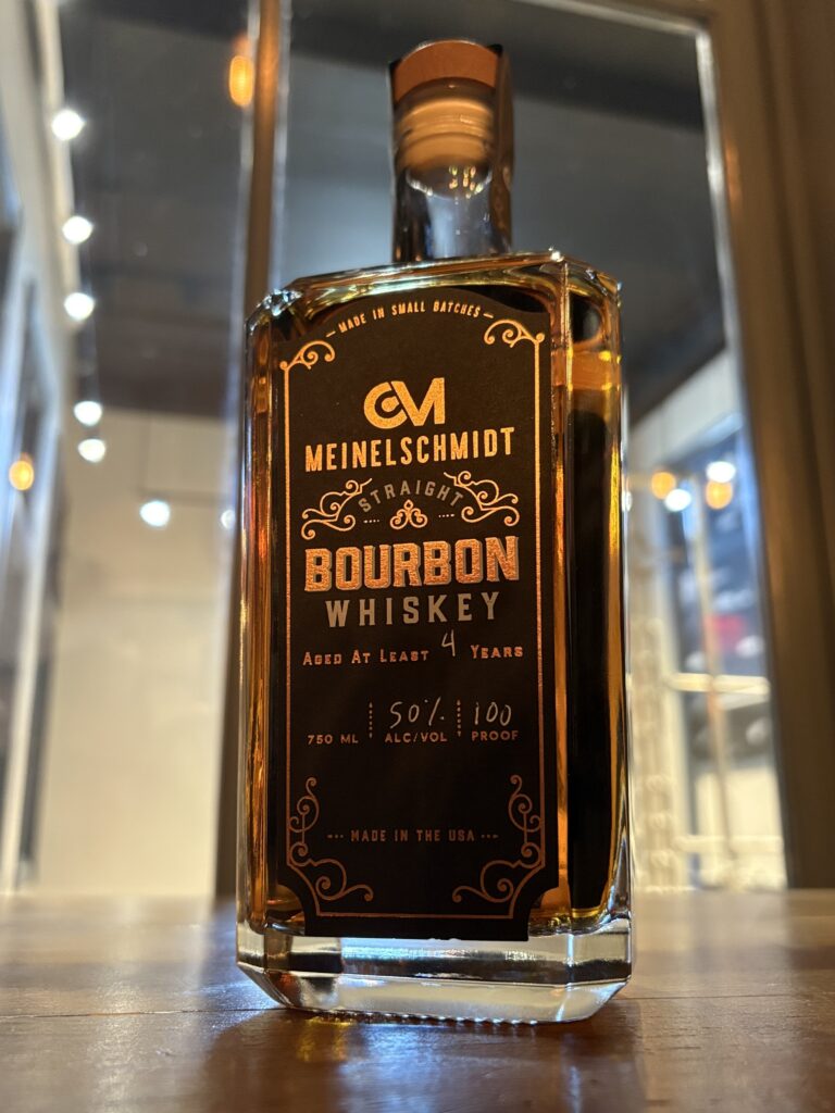 A bottle of Meinelschmidt Straight Bourbon Whiskey sits on a wooden surface with warm lighting and blurred background, showcasing its label and amber liquid.
