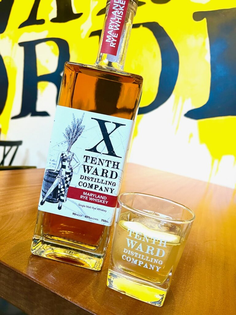 A bottle of Tenth Ward Maryland Rye Whiskey stands next to a glass with whiskey and ice, both on a wooden table, with a bold yellow and black mural in the background.