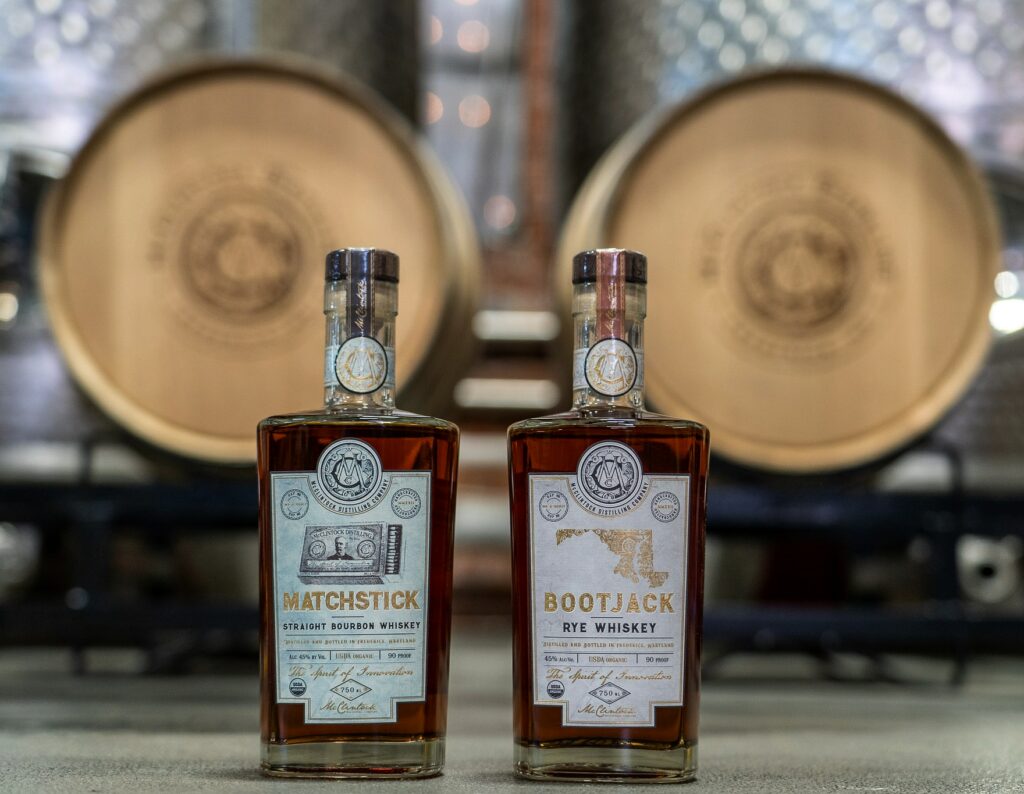 Two bottles of whiskey labeled Matchstick and Bootjack are displayed on a surface, with large wooden barrels blurred in the background inside a distillery.