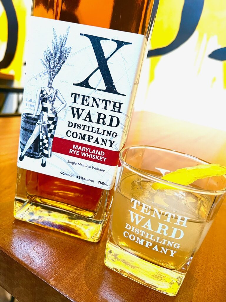 A bottle of Tenth Ward Distilling Company Maryland Rye Whiskey beside a glass with whiskey and a lemon twist, both on a wooden surface.