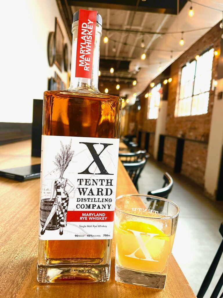 A bottle of Tenth Ward Maryland Rye Whiskey and a glass of whiskey sit on a wooden table in a rustic, brick-walled bar with warm lighting and empty chairs.