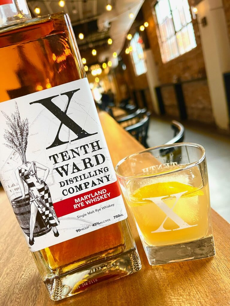 A bottle of Tenth Ward Maryland Rye Whiskey and a glass with whiskey and an orange peel sit on a wooden bar in a warmly lit, rustic setting.