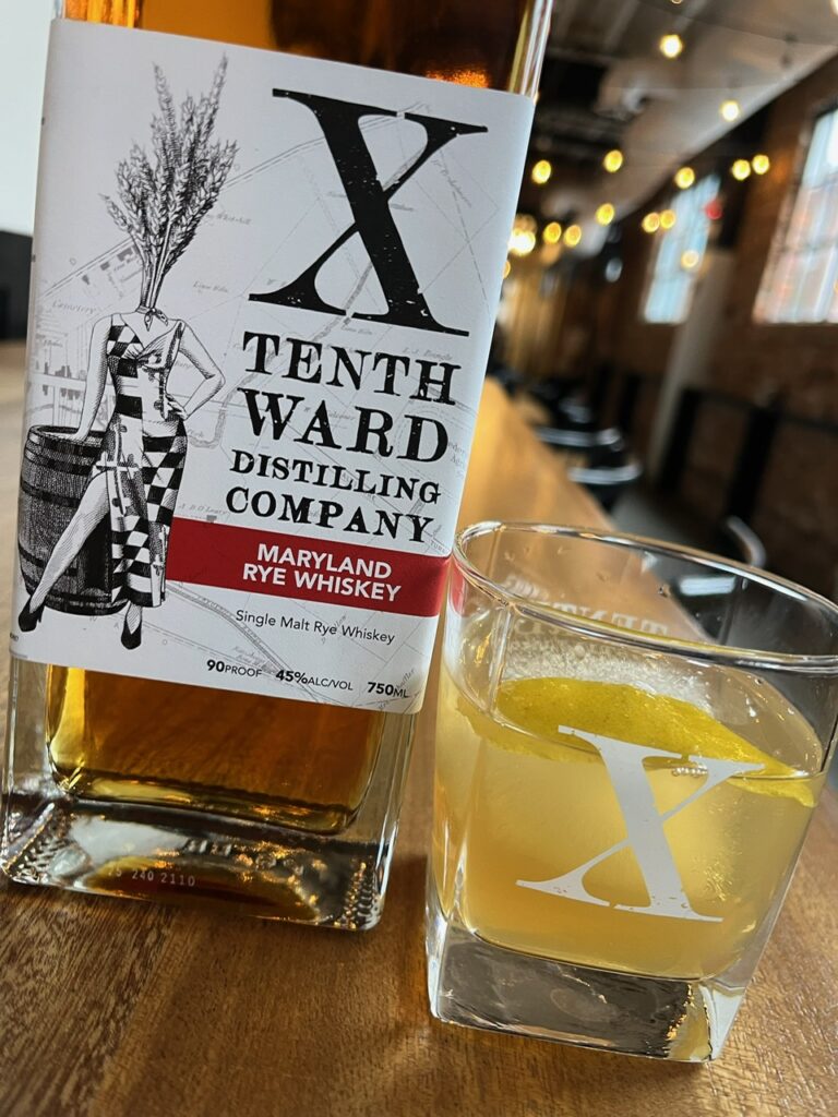 A bottle of Tenth Ward Distilling Company Maryland Rye Whiskey stands next to a glass of whiskey on a wooden bar, both featuring the distillery's “X” logo, with a blurred, brick-walled bar in the background.