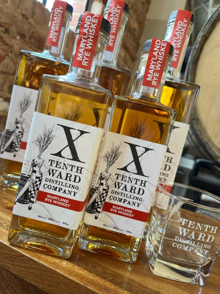 Three bottles of Tenth Ward Distilling Company Maryland Rye Whiskey and a branded glass are displayed on a wooden surface, with a whiskey barrel blurred in the background.