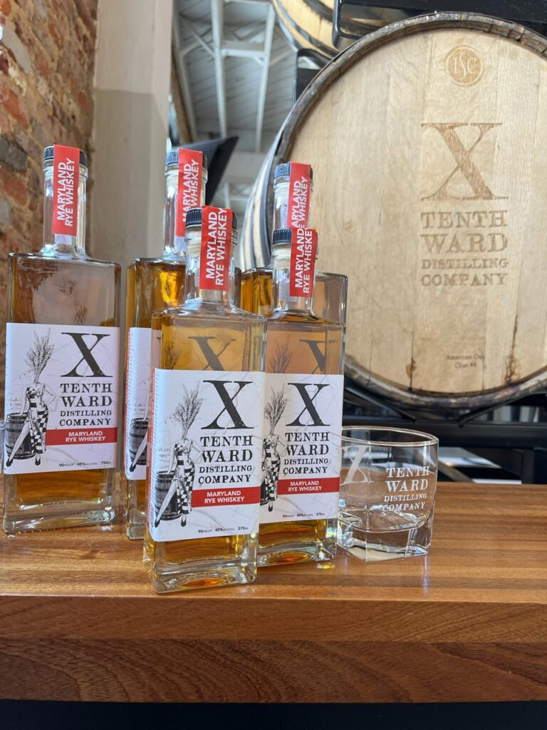 Four Tenth Ward Distilling Company rye whiskey bottles are displayed on a wooden table beside a branded glass and a large barrel marked with the company’s logo in the background.