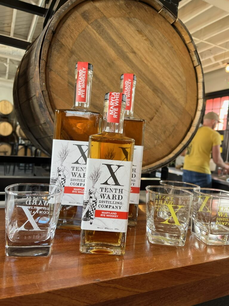 Three bottles of Tenth Ward Distilling Company rye whiskey sit on a bar with branded glasses in front; a large wooden barrel and person in a yellow shirt are visible in the background.