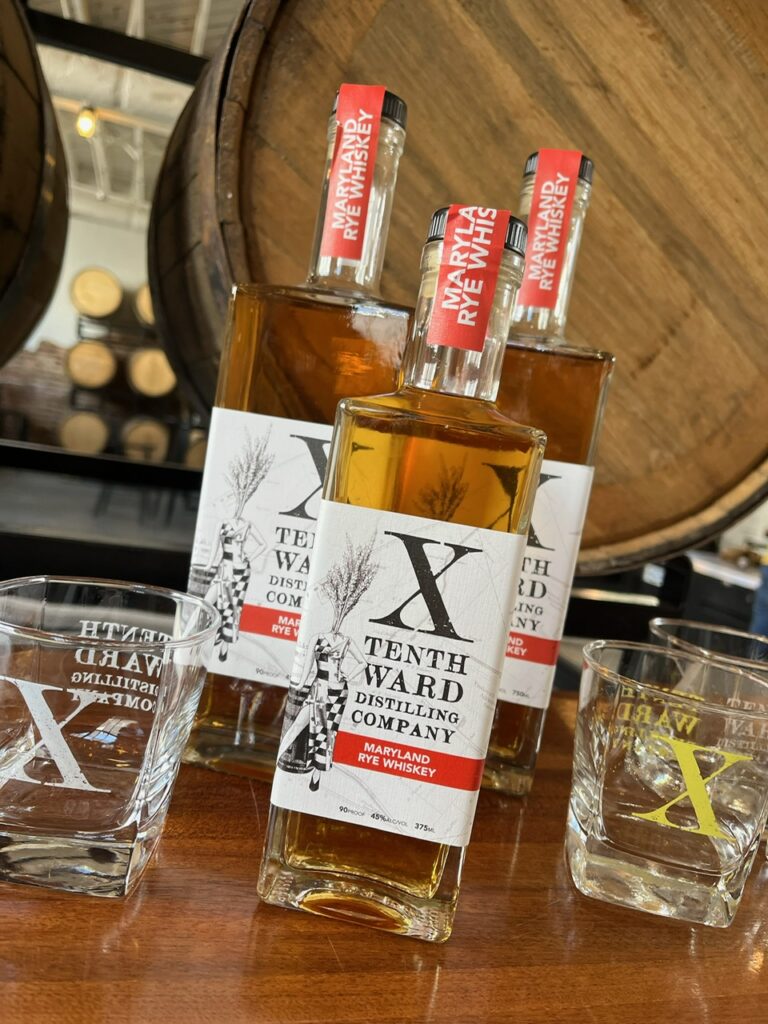 Three bottles of Tenth Ward Maryland Rye Whiskey and two branded glasses are displayed on a table in front of a large wooden barrel inside a distillery.