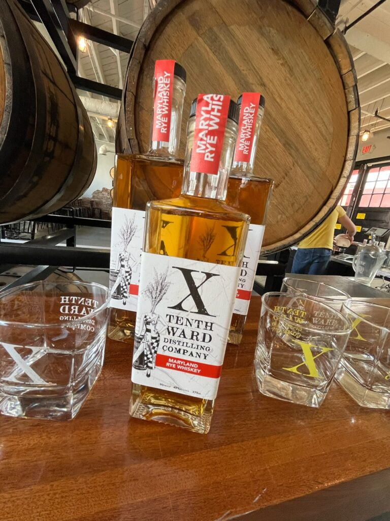 Three bottles of Tenth Ward rye whiskey are displayed on a table with four branded glasses in front, and large wooden barrels are visible in the background.