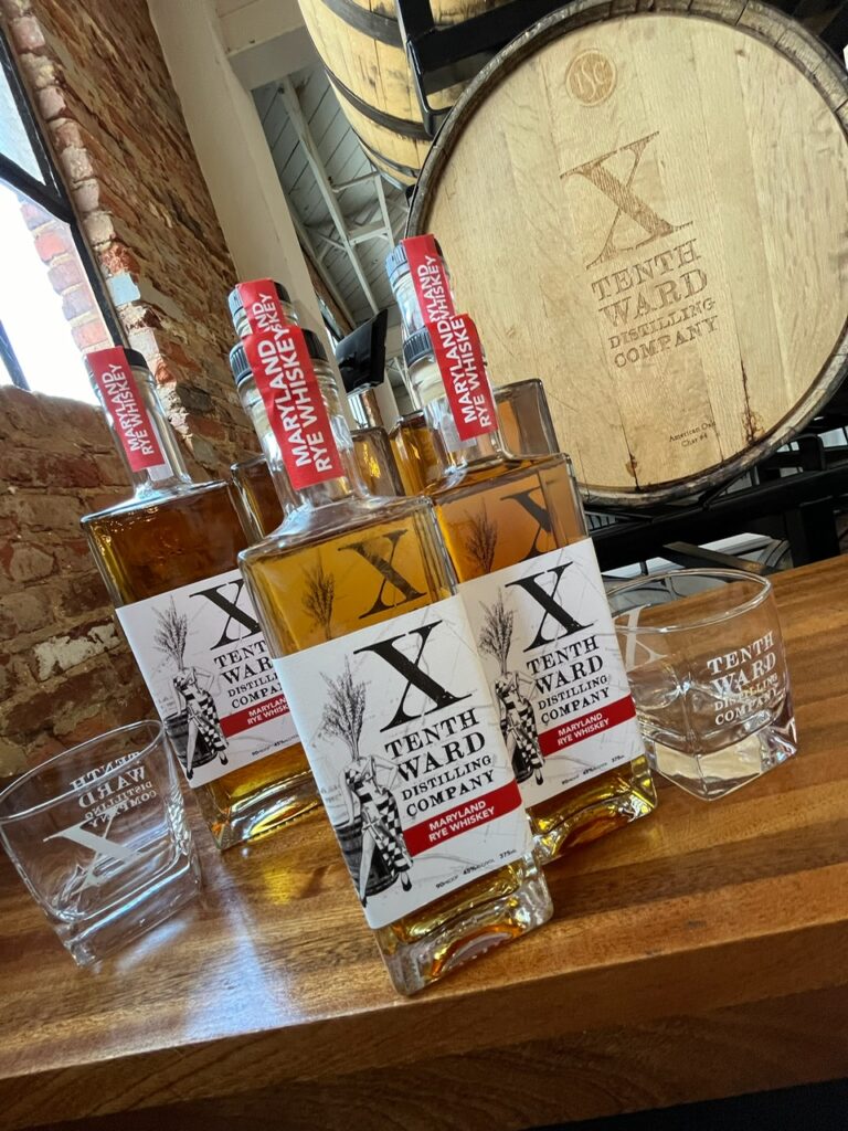 Three bottles of Tenth Ward rye whiskey, two glasses, and a large wooden barrel with the Tenth Ward logo are displayed on a wooden table in a distillery setting with brick walls and natural lighting.