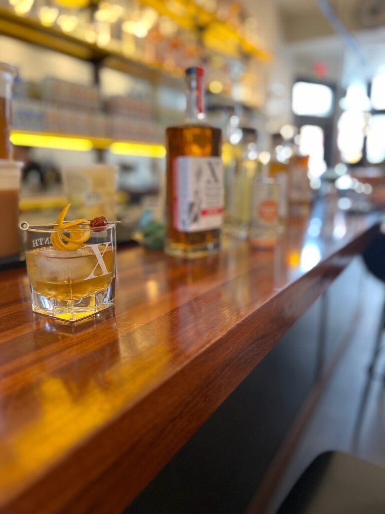 A glass of whiskey with an orange twist and cherry sits on a polished bar, surrounded by whiskey bottles. The background is softly blurred, with shelves and natural light visible.