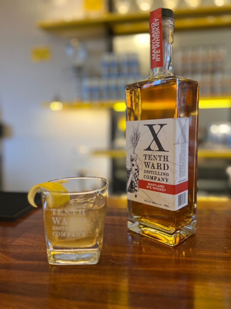 A bottle of Tenth Ward whiskey and a glass with whiskey, ice, and a lemon twist sit on a wooden bar, with shelves and bottles blurred in the background.