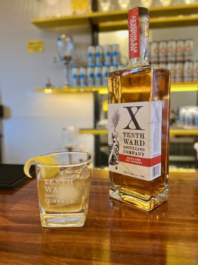 A bottle of Tenth Ward rye whiskey stands beside a glass of whiskey with ice and a lemon twist, both on a wooden bar with shelves and cans blurred in the background.