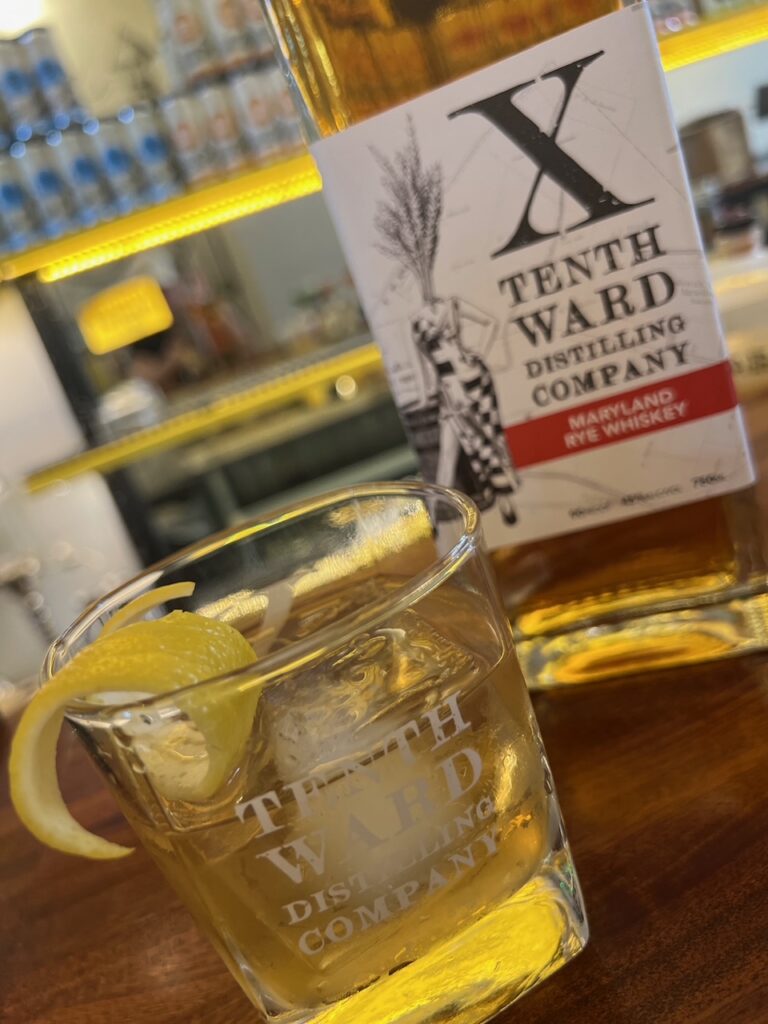 A glass with whiskey, ice, and a lemon twist sits on a bar next to a Tenth Ward Distilling Company Maryland rye whiskey bottle. Shelves with bottles are blurred in the background.