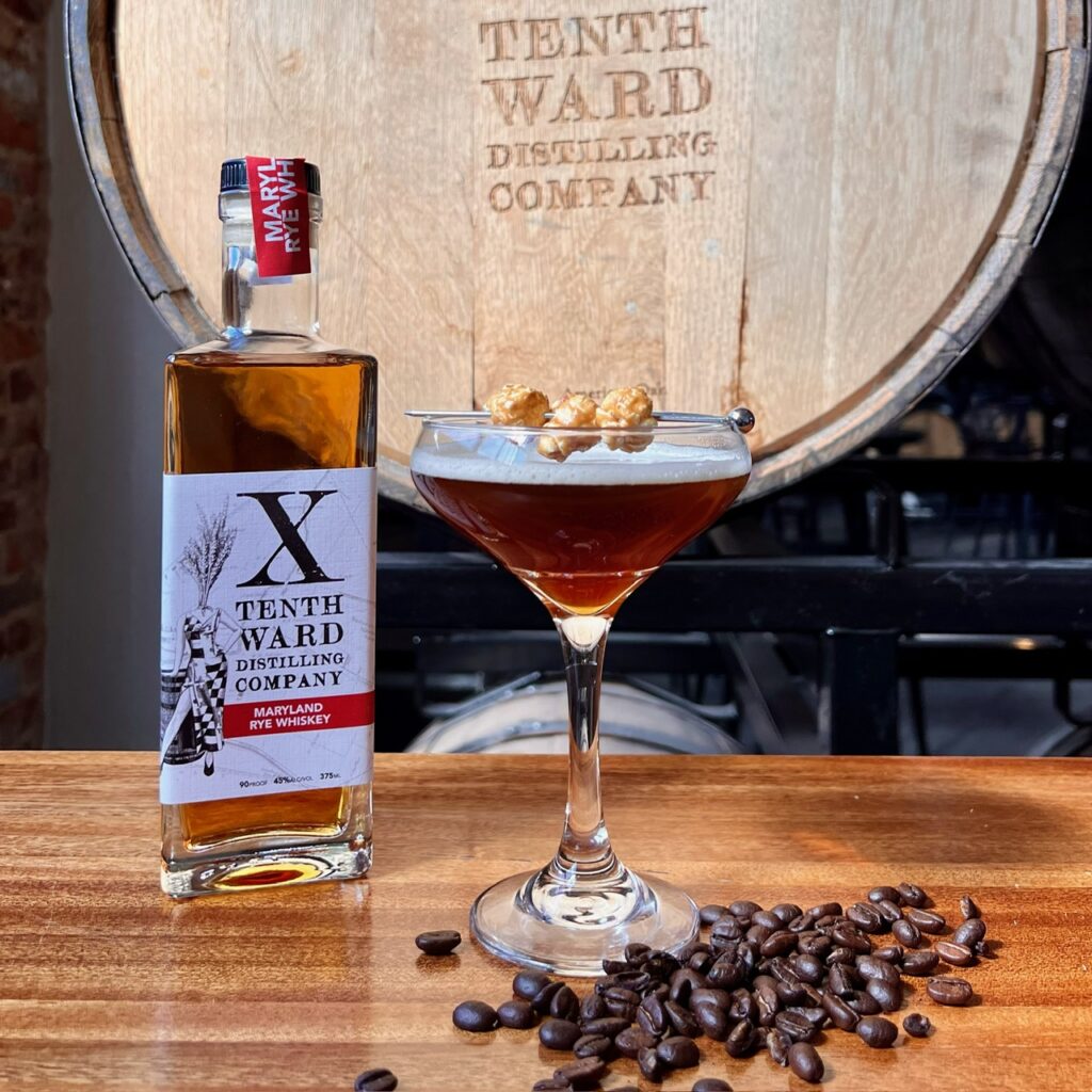 A bottle of Tenth Ward Maryland Rye Whiskey, a cocktail in a martini glass garnished with popcorn, and coffee beans are arranged on a table in front of a Tenth Ward Distilling Company barrel.
