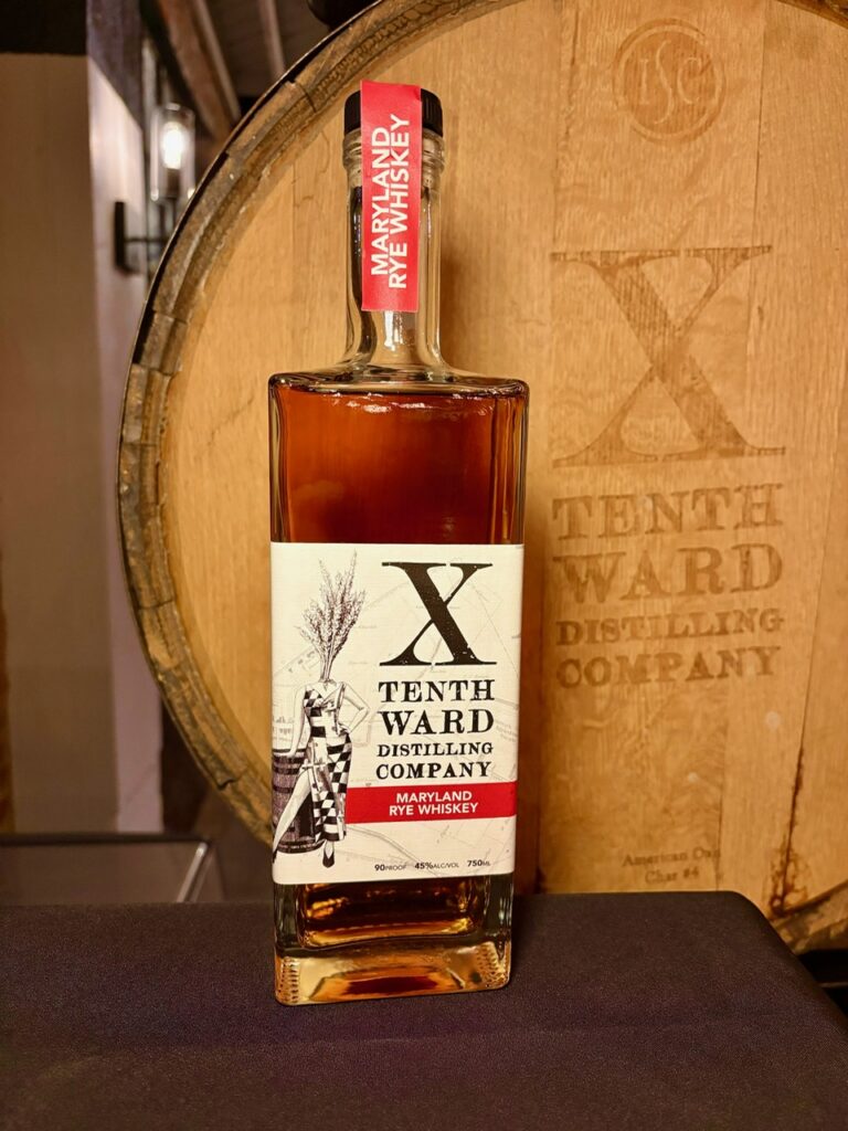 A bottle of Tenth Ward Distilling Company Maryland Rye Whiskey stands in front of a wooden barrel with the Tenth Ward logo and name engraved on it.