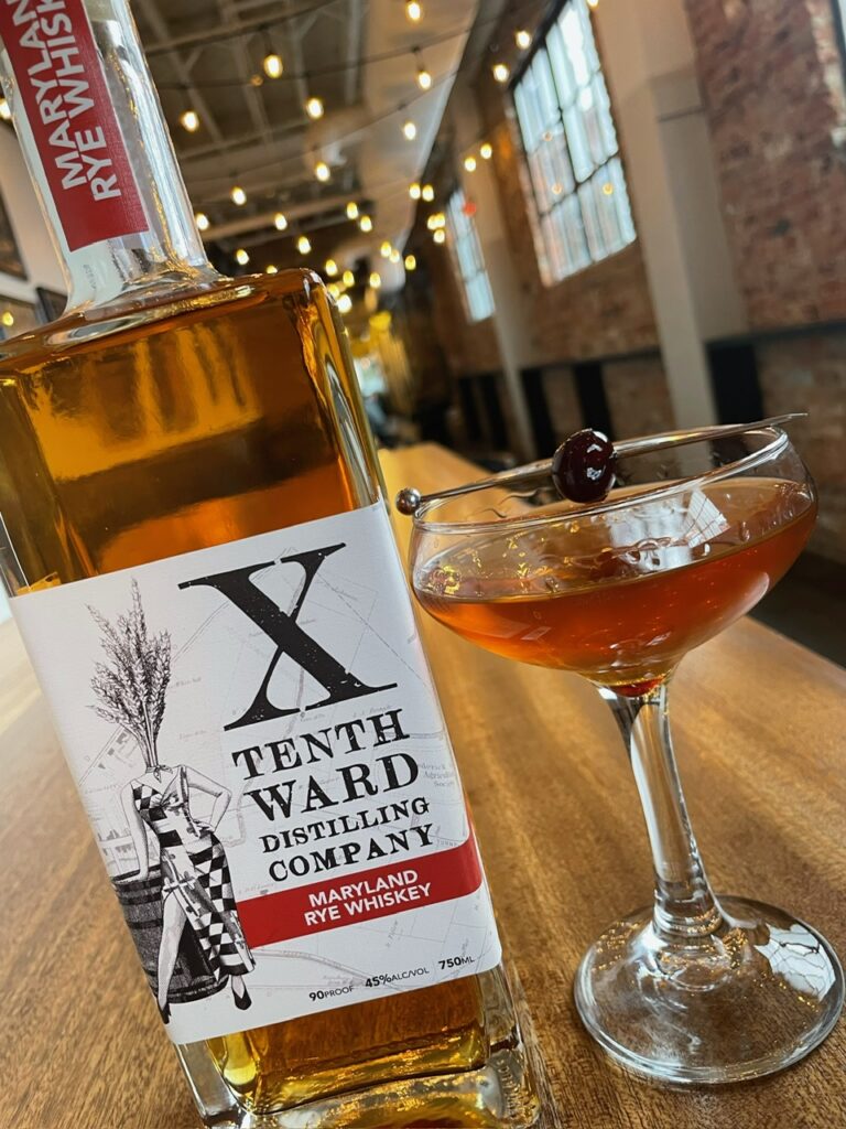 A bottle of Tenth Ward Maryland Rye Whiskey stands on a wooden table next to a cocktail garnished with a cherry, inside a cozy, well-lit bar with string lights and exposed brick walls.