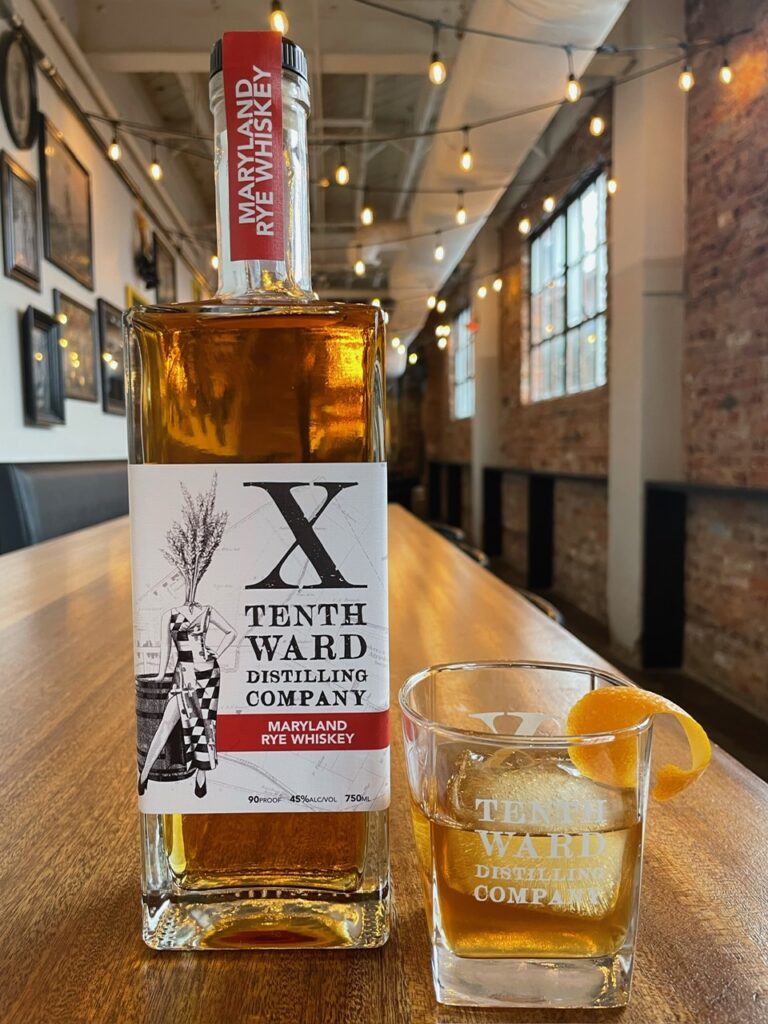 A bottle of Tenth Ward Maryland Rye Whiskey and a glass with whiskey, ice, and an orange twist sit on a wooden table in a cozy, well-lit bar with exposed brick walls and string lights.