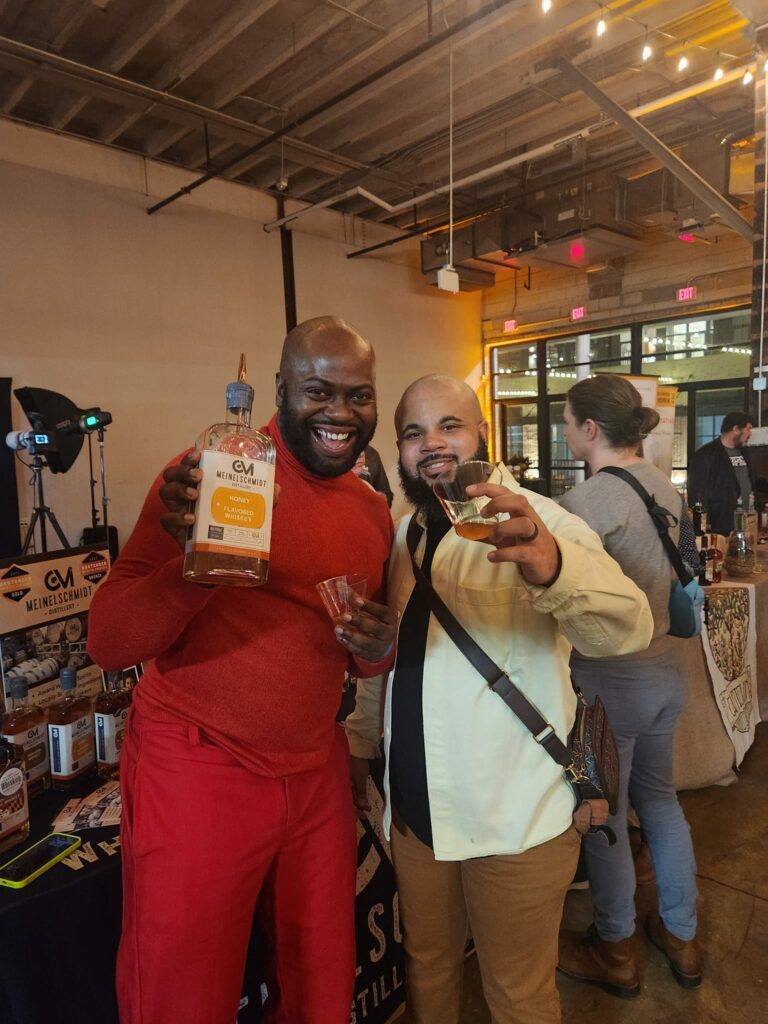 Two smiling men hold up drinks at an indoor event with tasting tables, bottles, and people in the background. One man wears red and holds a large bottle, while the other wears a cream shirt. Warm lighting fills the space.