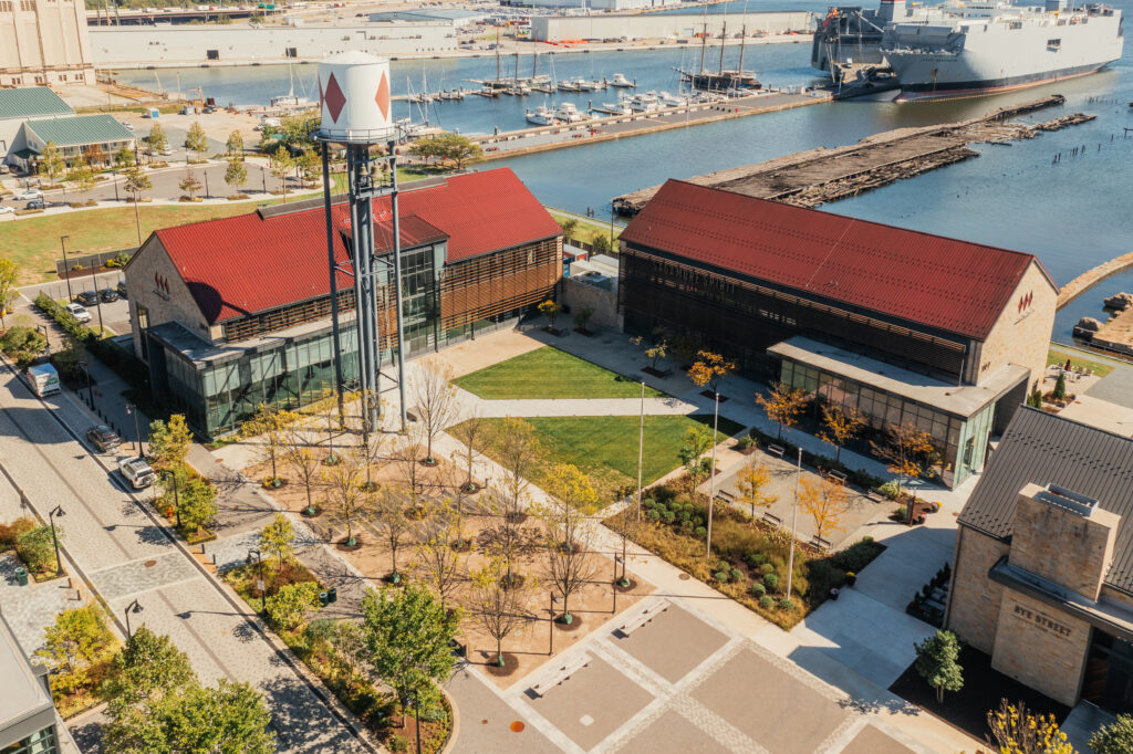 Aerial view of a waterfront area with two red-roofed buildings, a water tower, boats docked in a marina, landscaped paths, green lawns, and nearby industrial and commercial structures.