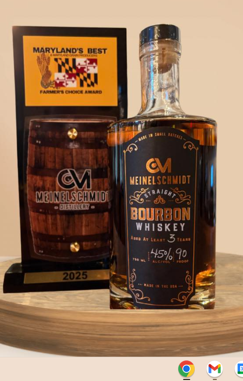 A bottle of Meinelschmidt Bourbon Whiskey stands next to a 2025 Maryland's Best Farmer's Choice Award trophy on a wooden surface. Computer screen icons are visible at the bottom right.