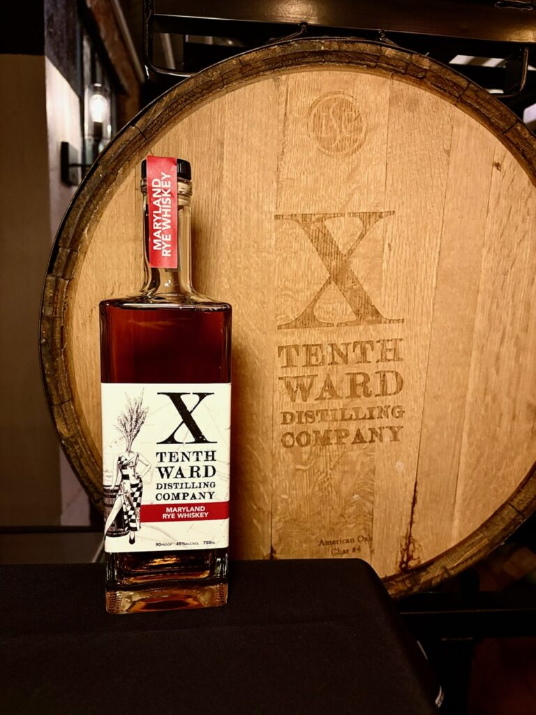 A bottle of Tenth Ward Distilling Company rye whiskey stands in front of a large wooden barrel engraved with the company’s name and logo.