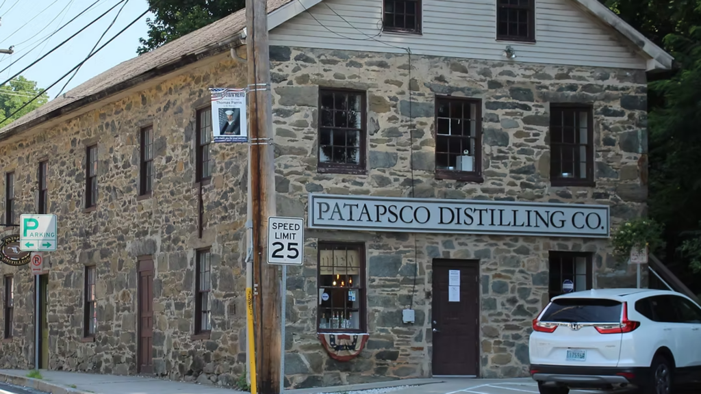 A stone building with a sign reading “Patapsco Distilling Co.” stands by the street. A white SUV is parked outside, and a speed limit 25 sign is visible nearby.