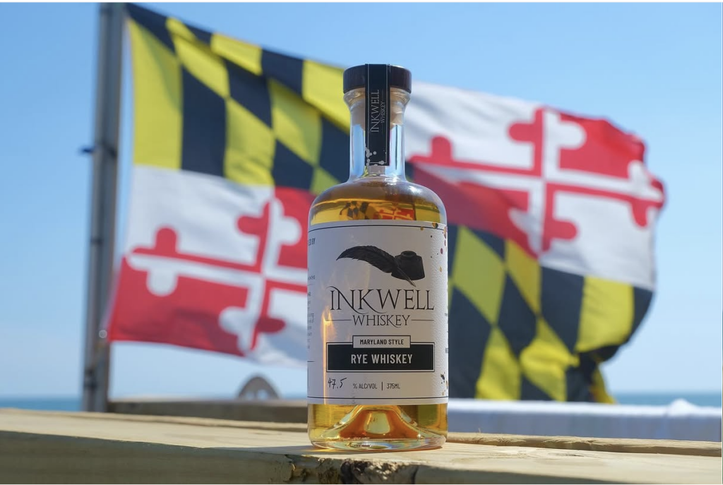 A bottle of Inkwell Rye Whiskey sits on a wooden surface with a Maryland state flag waving in the background on a sunny day.