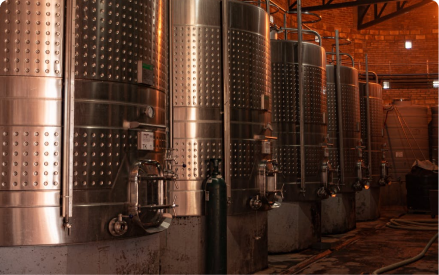Rows of large, stainless steel fermentation tanks in a winery or brewery, set in an industrial indoor space with brick walls and pipes running along the floor and ceiling.