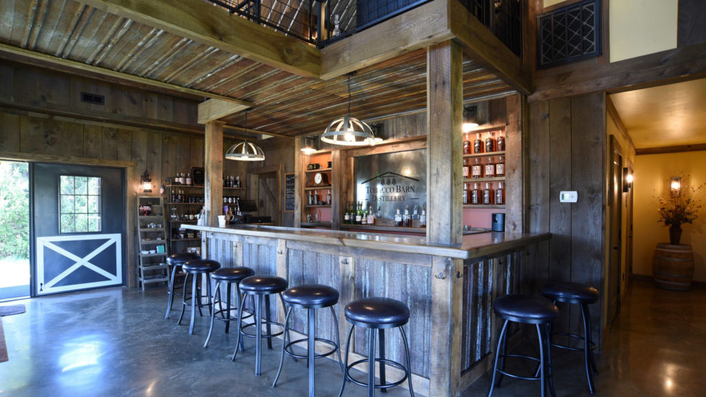 Rustic bar with wood paneling, metal stools, hanging lights, shelves of liquor, and a large mirror behind the counter. There is a loft above and a door with natural light entering on the left side.