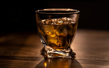 A glass of whiskey with ice cubes sits on a wooden surface, illuminated by warm, soft lighting against a dark background.