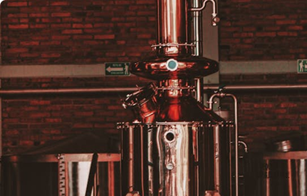 A shiny copper and metal distillation apparatus stands in front of a brick wall, surrounded by steel brewing tanks and pipes in an industrial setting.