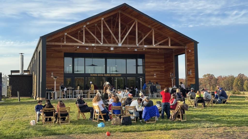 People sit on chairs outside a wooden building marked “Sowing Dog Farm Distillery,” enjoying a sunny day with music, drinks, and conversation on a grassy lawn.
