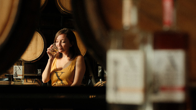 A woman in a yellow tank top sits among barrels and drinks from a glass, with bottles blurred in the foreground.