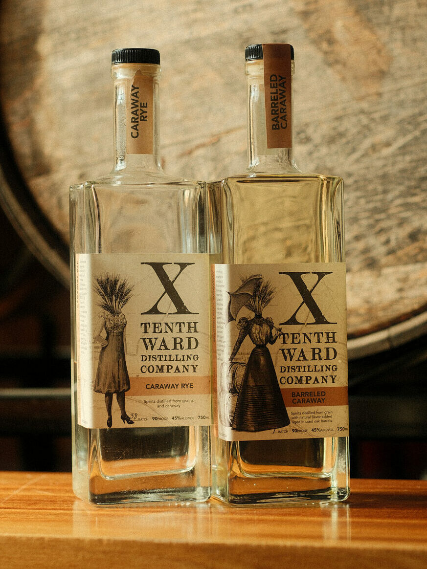 Two bottles of Tenth Ward Distilling Company spirits sit on a wooden surface, with a large barrel in the background. The labels feature vintage-style illustrations and text indicating Caraway Rye and Barreled Caraway Rye.